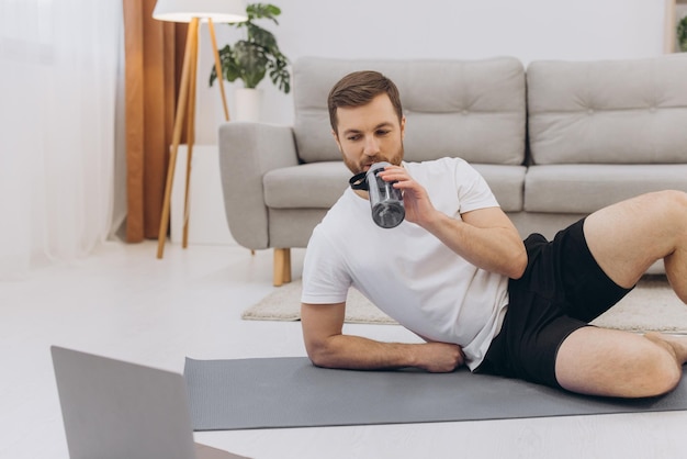 Schöner erwachsener Mann, der Laptop und Trinkwasser benutzt, während er zu Hause eine Pause während des Trainings hat