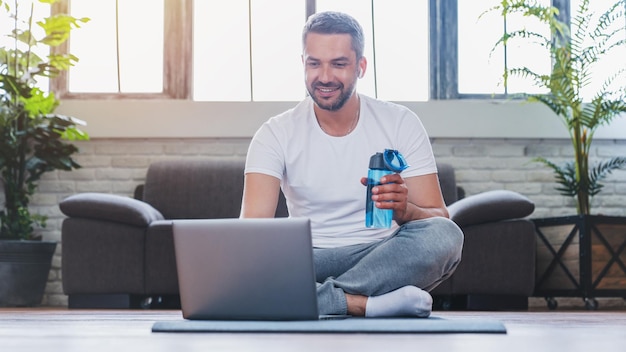 Schöner erwachsener Mann, der Laptop und Trinkwasser benutzt, während er zu Hause eine Pause während des Trainings hat