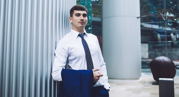 Schöner erwachsener Mann in eleganter Kleidung, der sich mit der Hand in der Tasche in der hellen Lobby des Bürogebäudes auf eine Metallsäule stützt und in die Kamera blickt