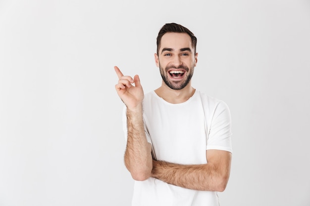 Schöner fröhlicher Mann mit leerem T-Shirt, das isoliert über weißer Wand steht und mit dem Finger auf den Kopierraum zeigt