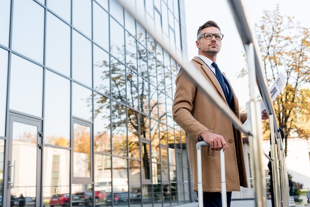 Schöner Geschäftsmann, der in beigem Mantel steht und Wirtschaftszeitung in der Nähe des Koffers hält