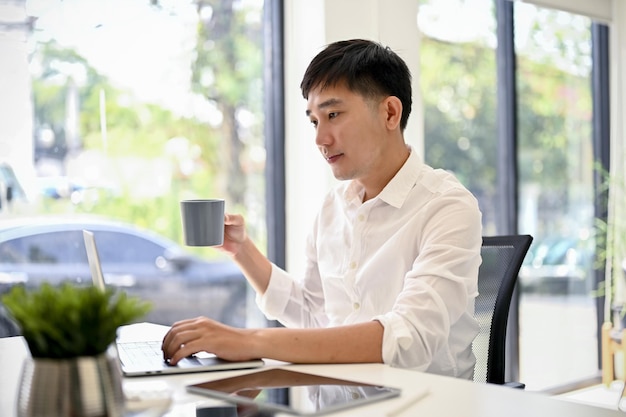 Schöner Geschäftsmann, der seinen Laptop benutzt und an seinem Schreibtisch in einem modernen Büro Kaffee trinkt