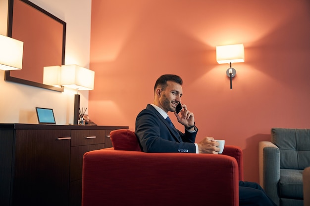 Schöner Geschäftsmann in einem Anzug, der sich bei einer Tasse Kaffee im Sessel entspannt und beim Telefonieren lächelt