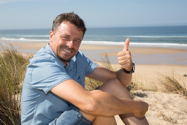 Schöner glücklicher Mann, der am Strand Daumen hochgibt