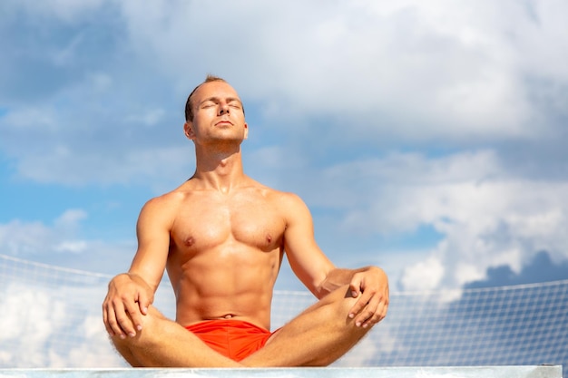 Schöner, hemdloser junger Mann während der Meditation oder bei einer Outdoor-Yoga-Übung, die gegen den blauen Himmel sitzt