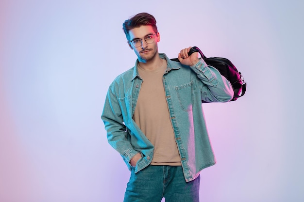 Foto schöner hipster-geschäftsmann mit stylischer brille in ausgefallener denim-kleidung und einem jeanshemd mit einer modischen ledertasche in einem studio mit kreativer farbiger rosa und blauer kommerzieller beleuchtung
