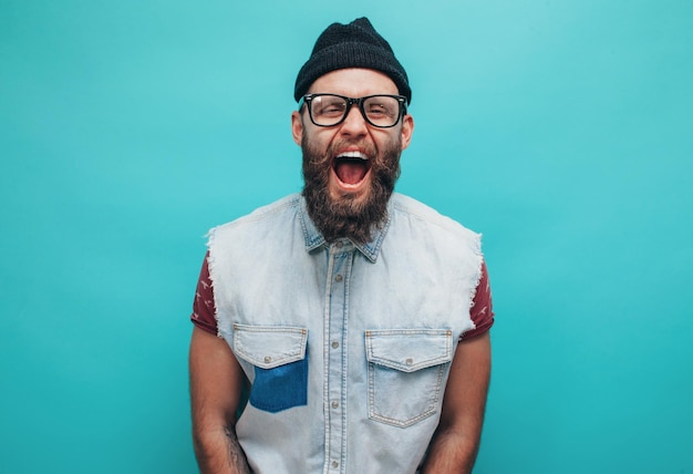 Foto schöner hipster-typ mit bart, der glücklich lächelt, lässig gekleidet mit schwarzer mütze auf dem kopf, isoliert über einem blauen studiohintergrund das konzept der verrückten emotionen der menschen