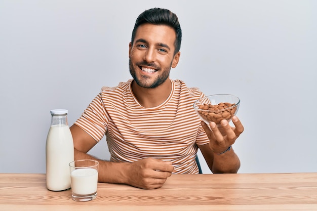 Schöner hispanischer Mann, der gesunde Mandelmilch trinkt, sieht positiv und glücklich aus und lächelt mit einem selbstbewussten Lächeln, das Zähne zeigt