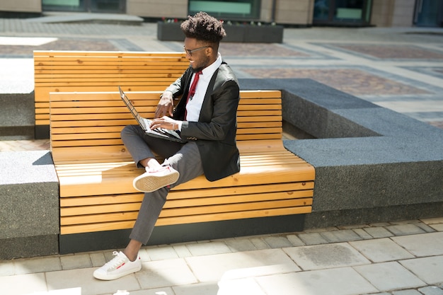 schöner junger afrikanischer Mann auf Bank mit Laptop im Sommer