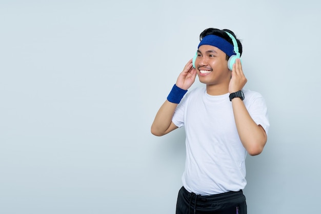 Schöner junger asiatischer Mann, sportlicher Fitnesstrainer-Lehrer in blauem Stirnband und weißem T-Shirt Macht Spaß, Musik mit Kopfhörern zu hören, die über weißem Hintergrund isoliert sind