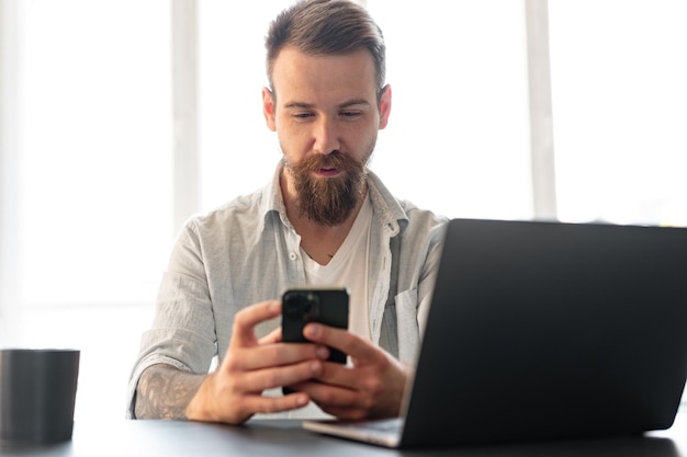 Schöner junger bärtiger Mann, der Zeit zu Hause mit dem Telefon in den Händen verbringt