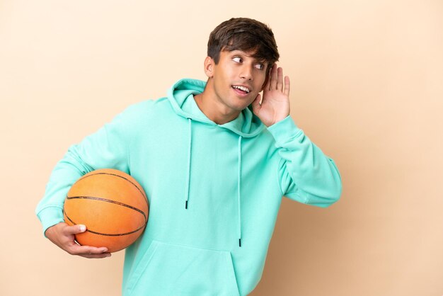 Schöner junger Basketballspieler isoliert auf ockerfarbenem Hintergrund, der etwas hört, indem er die Hand auf das Ohr legt