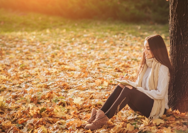 Schöner junger Brunette, der auf einem gefallenen Herbstlaub in einem Park sitzt, ein Buch liest oder ein Tagebuch schreibt