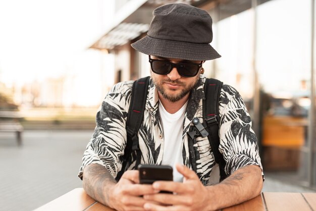 Schöner junger Hipster-Mann in modischer Kleidung mit Hut sitzt in einem Sommercafé und kommuniziert Surfen auf einem Smartphone Kerl mit Telefon reist in der Stadt