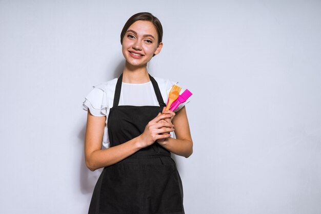 Schöner junger Mädchenkoch in schwarzer Schürze lächelt, hält Geräte zum Kochen und Backen in den Händen