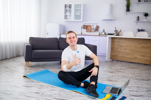 Schöner junger Mann, der auf dem Boden sitzt und eine Flasche Wasser hält, während er online trainiert