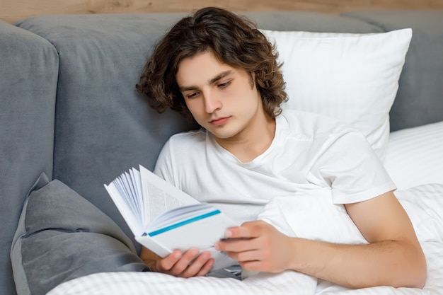 Schöner junger Mann, der auf seinem Bett liegt und ein Buch liest und sich in seinem Schlafzimmer ausruht