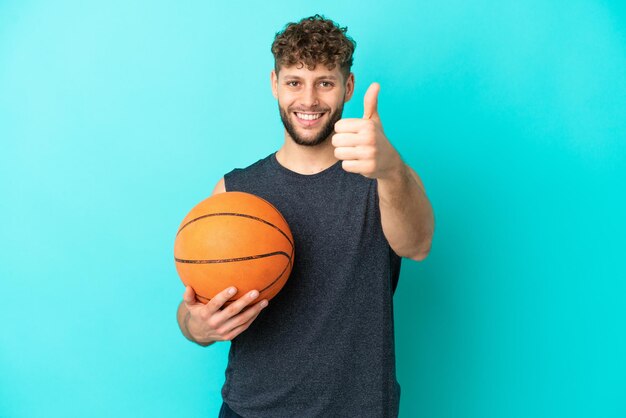 Schöner junger Mann, der Basketball isoliert auf blauem Hintergrund mit Daumen nach oben spielt, weil etwas Gutes passiert ist