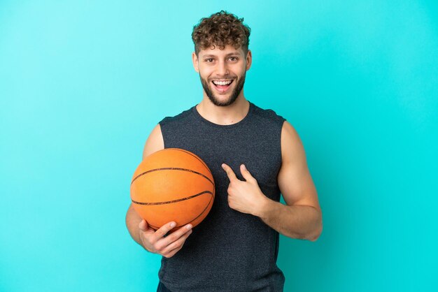 Schöner junger Mann, der Basketball spielt, isoliert auf blauem Hintergrund mit überraschendem Gesichtsausdruck