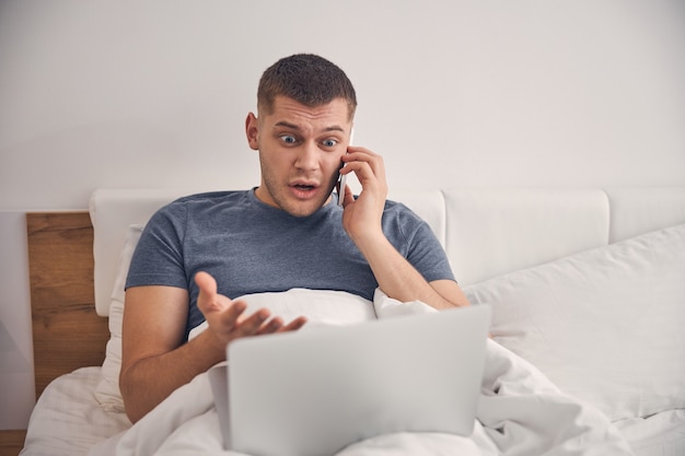 Schöner junger Mann, der im Bett am Laptop arbeitet, während er emotional mit verängstigtem Gesicht telefoniert