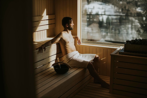 Schöner junger Mann, der sich in der Sauna entspannt und den Winterwald durch das Fenster beobachtet