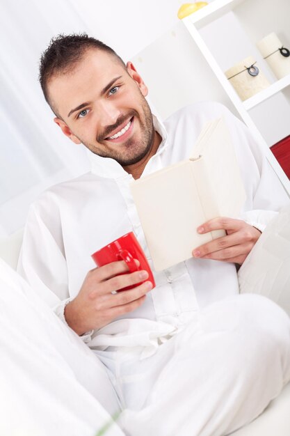 Schöner junger Mann, der sich mit Buch ausruht und zu Hause Kaffee trinkt.