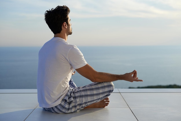 Schöner junger Mann, der Yoga auf der modernen Hausterrasse mit Meer und Sonnenuntergang im Hintergrund praktiziert
