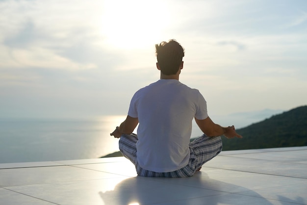 Schöner junger Mann, der Yoga auf der modernen Hausterrasse mit Meer und Sonnenuntergang im Hintergrund praktiziert