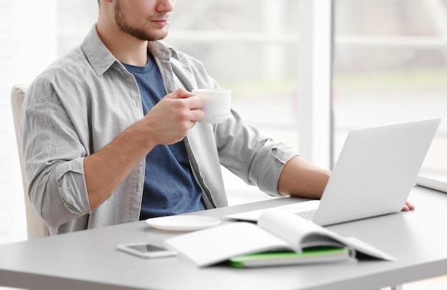 Schöner junger Mann, der zu Hause am Laptop arbeitet und Kaffee trinkt