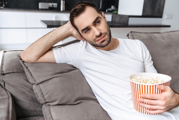 Schöner junger Mann, der zu Hause auf einer Couch sitzt, Popcorn isst, fernsieht