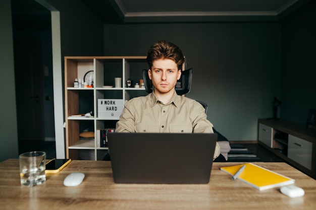 Schöner junger Mann in einem Hemd, der in einer gemütlichen Wohnung sitzt und an einem Laptop arbeitet, der mit ernstem Gesicht in die Kamera schaut Freiberufler arbeitet zu Hause in einem gemütlichen Raum Quarantäne