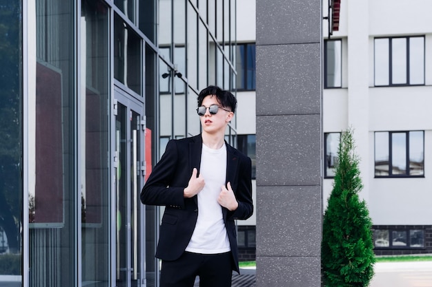 Schöner junger Mann in einem schwarzen Hemd, der vor dem Hintergrund von Stadtgebäuden steht und durch eine schwarze Sonnenbrille wegschaut