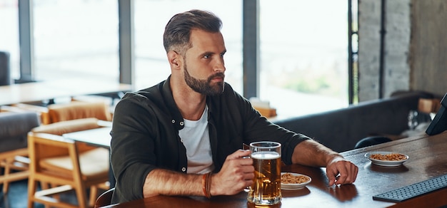 Schöner junger Mann in Freizeitkleidung, der Bier genießt, während er Zeit in der Kneipe verbringt?