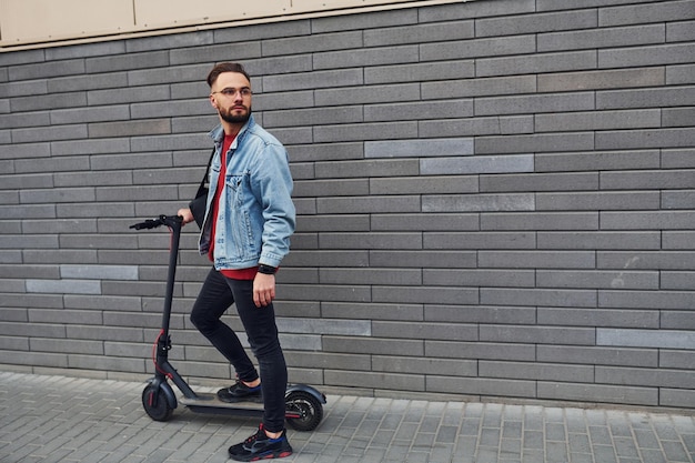 Schöner junger Mann in Freizeitkleidung, der tagsüber mit Elektroroller im Freien steht