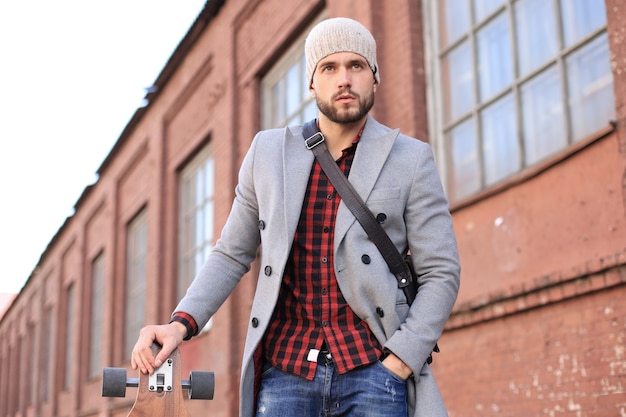 Schöner junger Mann in grauem Mantel und Hut, der mit Longboard auf der Straße geht.