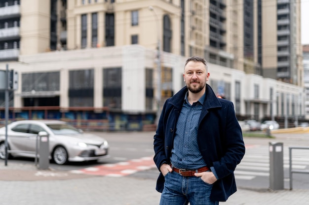 Schöner junger Mann in grauem Mantel und Jeans, der beim Überqueren der Straße vor der Kamera posiert. Foto in halber Länge. Nahansicht.