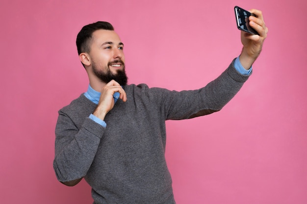 Schöner junger Mann in lässiger, stilvoller Kleidung, der isoliert über der Hintergrundwand steht und ein Smartphone hält, das ein Selfie-Foto mit Blick auf das Display des Mobiltelefons macht