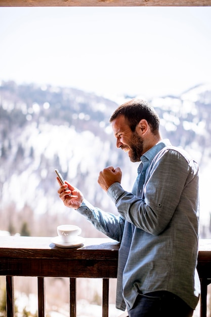 Schöner junger Mann mit Handy mit Tasse heißen Tee am Winterfenster