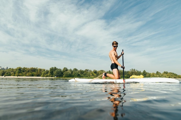 Schöner junger Mann mit Sporttorso paddelt ruhig auf einem Sup-Board auf dem Fluss