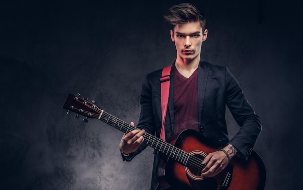 Schöner junger Musiker mit stylischem Haar in eleganter Kleidung mit einer Gitarre in den Händen, die auf dunklem Hintergrund spielt und posiert.