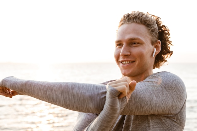 Schöner junger Sportler, der am Strand steht und Musik mit Kopfhörern hört, macht Dehnübungen.