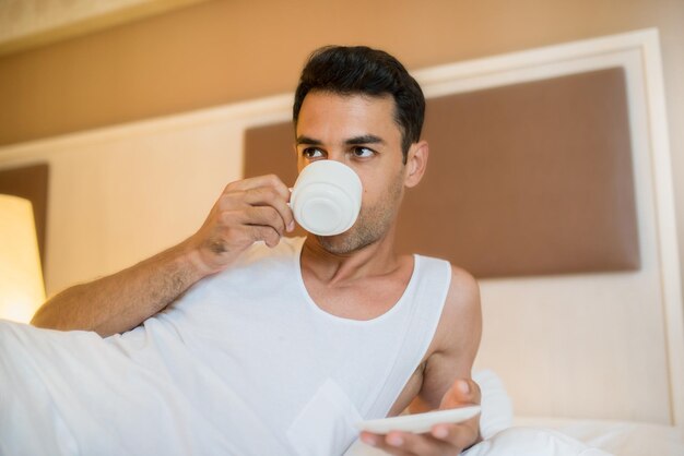 Schöner junger wacher Mann, der eine Tasse Kaffee im weißen Bett trinkt Schlafzimmerhintergrund