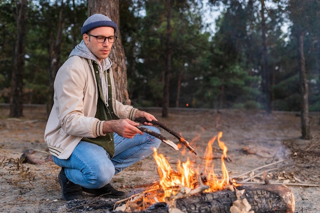 Schöner kaukasischer Mann in warmer Kleidung, der mit Stöcken Feuer macht, während er in der Nähe von trockenen Ästen sitzt. Wollen Sie warmes Konzept halten