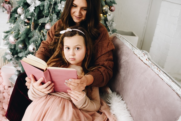 Schöner kaukasischer Moher, der mit ihrer reizenden kleinen Tochter sitzt, die lila Buch nahe geschmücktem Weihnachtsbaum liest