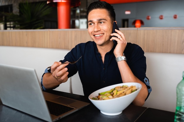 Foto schöner kerl, der mit lächelndem gesicht am handy spricht, während er salat mit dem computer isst