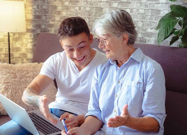 Schöner kleiner Junge, der zu Hause auf dem Sofa sitzt und der älteren Großmutter hilft, mit dem Laptop im Internet zu surfen, während sie sich Notizen macht. Jüngere Generationen kümmern sich um ältere Verwandte, die ihnen beibringen, wie man Computer benutzt