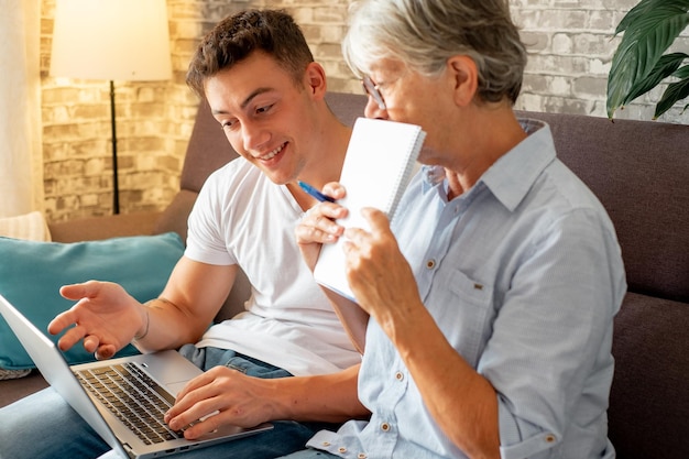 Schöner kleiner Junge, der zu Hause auf dem Sofa sitzt und der älteren Großmutter hilft, mit dem Laptop im Internet zu surfen, während sie sich Notizen macht. Jüngere Generationen kümmern sich um ältere Verwandte, die ihnen beibringen, wie man Computer benutzt