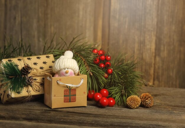 Schöner kleiner Schneemann im Paket, das mit einem anderen Geschenk auf dem Holzhintergrund liegt, Tannenzweige, Zapfen und Holztannen sind in der Nähe