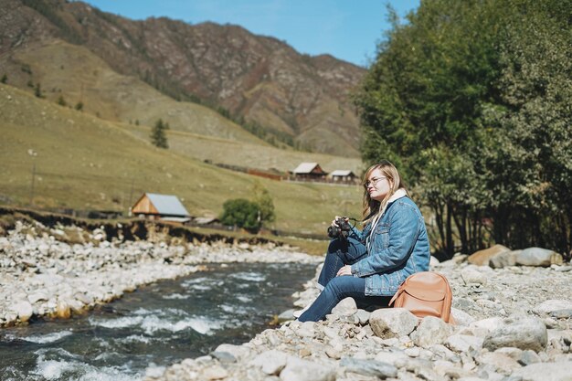 Schöner lächelnder blonder Reisender der jungen Frau mit Fotokamera in den Händen nahe dem Gebirgsfluss