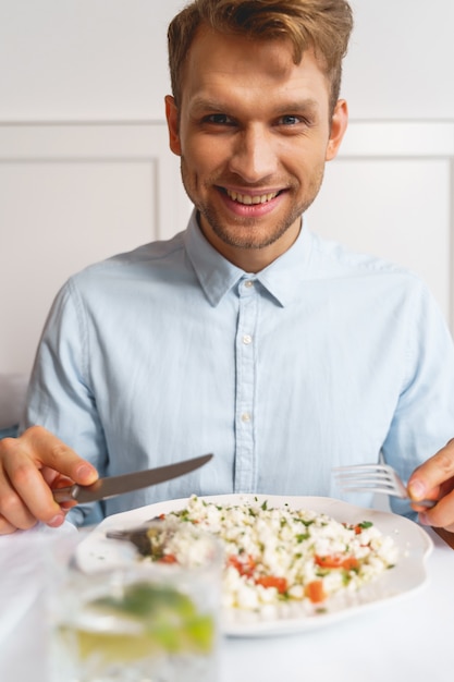 Foto schöner lächelnder herr, der gabel und messer hält, während er mit frischem salat am tisch sitzt sitting